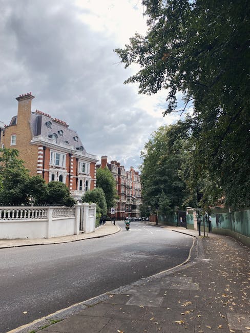 A street scene on Campden Hill Road in Holland Park showing a residential property with classic London-style architecture, including a multi-story brick building with white window frames, a slate roof, and decorative stonework. In the foreground, a black removal van belonging to Removal Van Holland Park is parked on the curb, with the side door open and furniture and cardboard boxes partially visible inside. Nearby, several wrapped and packed items, possibly furniture pieces and boxes, are being prepared for loading onto the van. The area is shaded by large leafy trees, with some fallen autumn leaves visible on the pavement, and a person can be seen near the items, assisting with the loading process. The scene captures the logistics of home relocation, with careful handling of packing materials and furniture as part of a professional removals service in a residential neighbourhood.