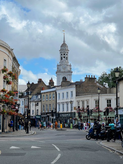 A street scene near a historic town centre features a large clock tower with a weather vane on top, situated behind a row of multi-storey traditional buildings with white and beige facades. In the foreground, there is a paved road with white markings, and a pedestrian crossing visible. Pedestrians are walking along the sidewalks, some waiting at the crossing, while others are shopping or chatting near shopfronts, including a café and retail stores. Several parked vehicles, including bicycles and motorbikes, are along the street, with a few people loading or unloading items. The image captures a partly cloudy sky with patches of blue, and decorative hanging flower baskets are attached to black lamp posts lining the street. This scene reflects a busy urban area suitable for professional removal and home relocation services, as provided by Removal Van Holland Park, demonstrating the typical environment encountered during furniture transport and packing processes.