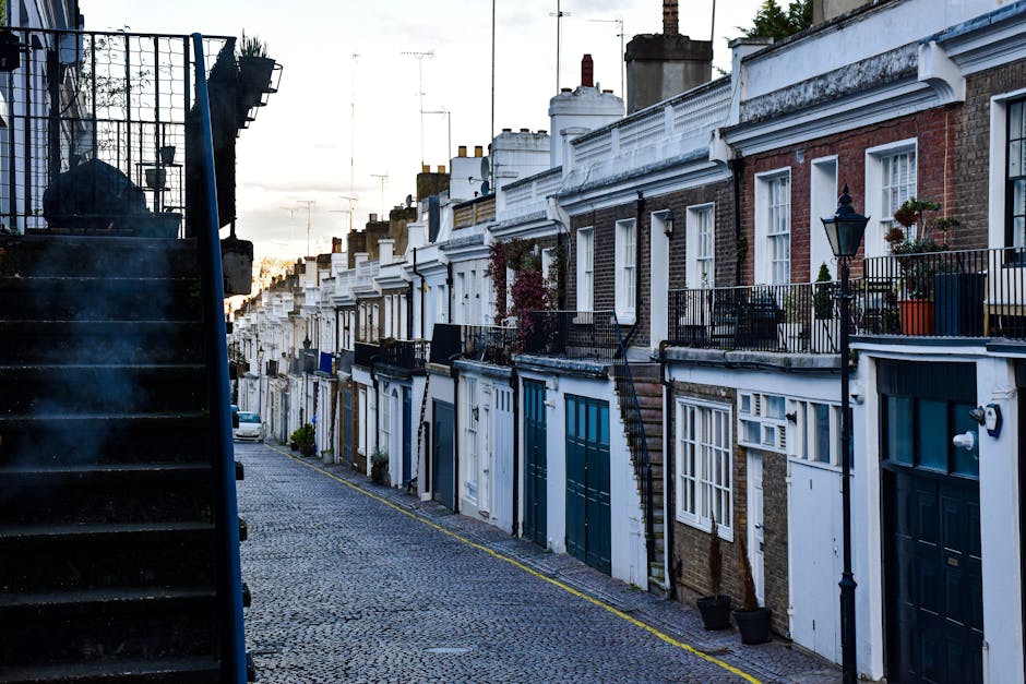 A street scene on Campden Hill Road in Holland Park showing a residential property with classic London-style architecture, including a multi-story brick building with white window frames, a slate roof, and decorative stonework. In the foreground, a black removal van belonging to Removal Van Holland Park is parked on the curb, with the side door open and furniture and cardboard boxes partially visible inside. Nearby, several wrapped and packed items, possibly furniture pieces and boxes, are being prepared for loading onto the van. The area is shaded by large leafy trees, with some fallen autumn leaves visible on the pavement, and a person can be seen near the items, assisting with the loading process. The scene captures the logistics of home relocation, with careful handling of packing materials and furniture as part of a professional removals service in a residential neighbourhood.