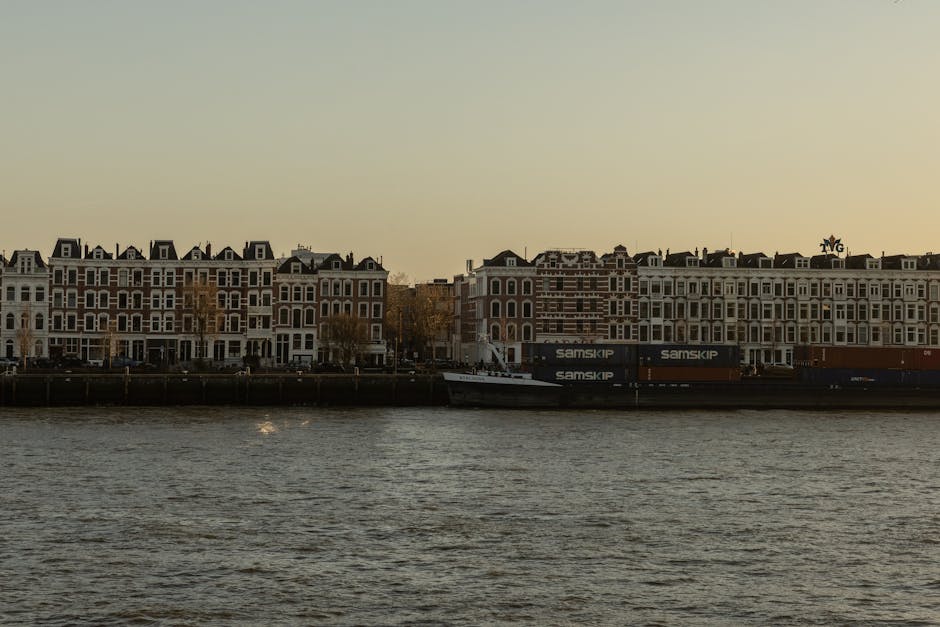 A row of multi-storey residential buildings along the riverbank in Holland Park Avenue, with large windows and brick facades, situated behind a paved walkway and a metal railing. Parked directly in fr