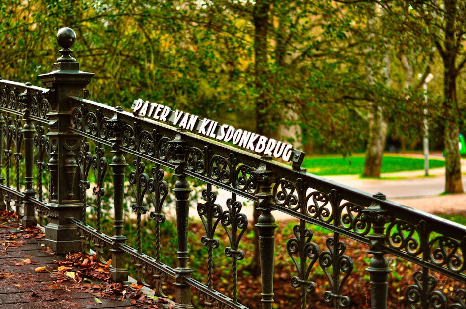 A close-up view of a decorative black wrought iron fence with intricate scrollwork, situated outdoors along a pathway covered in fallen autumn leaves. Attached to the fence is a white sign reading 'PA