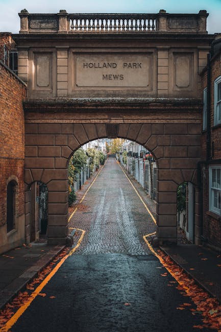 A view through the historic archway of Holland Park Mews, constructed from brown stone with an engraved inscription, leading onto a narrow cobbled street lined with wooden picket fences and residential houses. The street features a slight incline and is bordered by yellow lines along the edges, with fallen autumn leaves scattered on the wet pavement. In the foreground, the image captures part of a moving process with a removal van from Removal Van Holland Park parked nearby, loaded with stacks of cardboard boxes wrapped in plastic, furniture blankets, and a trolley, indicating ongoing house relocation activities. The scene is illuminated by natural daylight, highlighting the detailed textures of the stone arch, the cobblestones, and the packed items, illustrating elements typical of a home removal and furniture transport operation in the Holland Park area.
