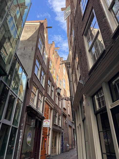 A narrow alleyway between tall, multi-storey brick buildings with various windows, some with open shutters and others closed, situated underneath a partly cloudy blue sky. On the right side, a white building with a small window is visible, while on the left, a modern glass-fronted structure reflects the surrounding architecture. A traditional black street lamp extends from the wall of one building, illuminating the area. The alley is relatively clean with a paved surface, and a small brown door with glass panels is seen at the ground level. The tight space illustrates the challenging access often encountered during home relocations or furniture transport, a scenario where removals specialist Removal Van Holland Park might be engaged to assist with narrow access remodelling and moving logistics in Holland Park.